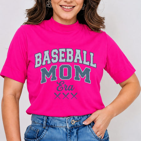 Woman wearing a bright hot pink mock neck t-shirt featuring a blue and white vintage-style "Baseball Mom Era" graphic print with crossed bats, tucked into blue jeans.