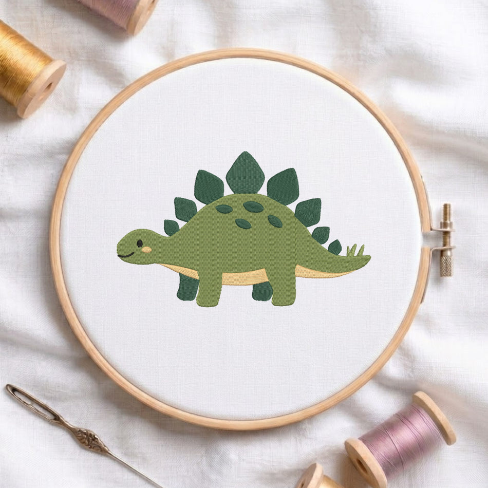 A wooden embroidery hoop holds a white fabric with a green dinosaur design in progress. The hoop is photographed on a white cloth with spools of thread and a needle around it.