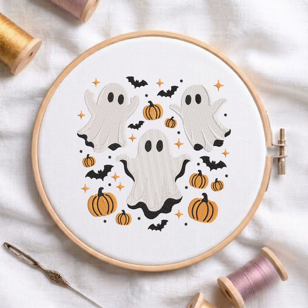 A white fabric stretched in a wooden embroidery hoop, featuring an embroidered Halloween scene with three cute ghosts, orange pumpkins, flying bats, and twinkling stars.