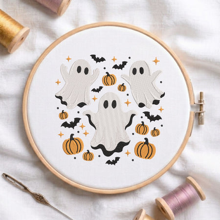 A white fabric stretched in a wooden embroidery hoop, featuring an embroidered Halloween scene with three cute ghosts, orange pumpkins, flying bats, and twinkling stars.
