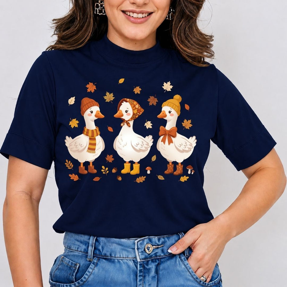 A woman wearing a navy blue short-sleeve t-shirt with a graphic of three white ducks dressed in autumn hats and scarves, surrounded by falling maple leaves. She is wearing blue jeans against a plain light gray background.