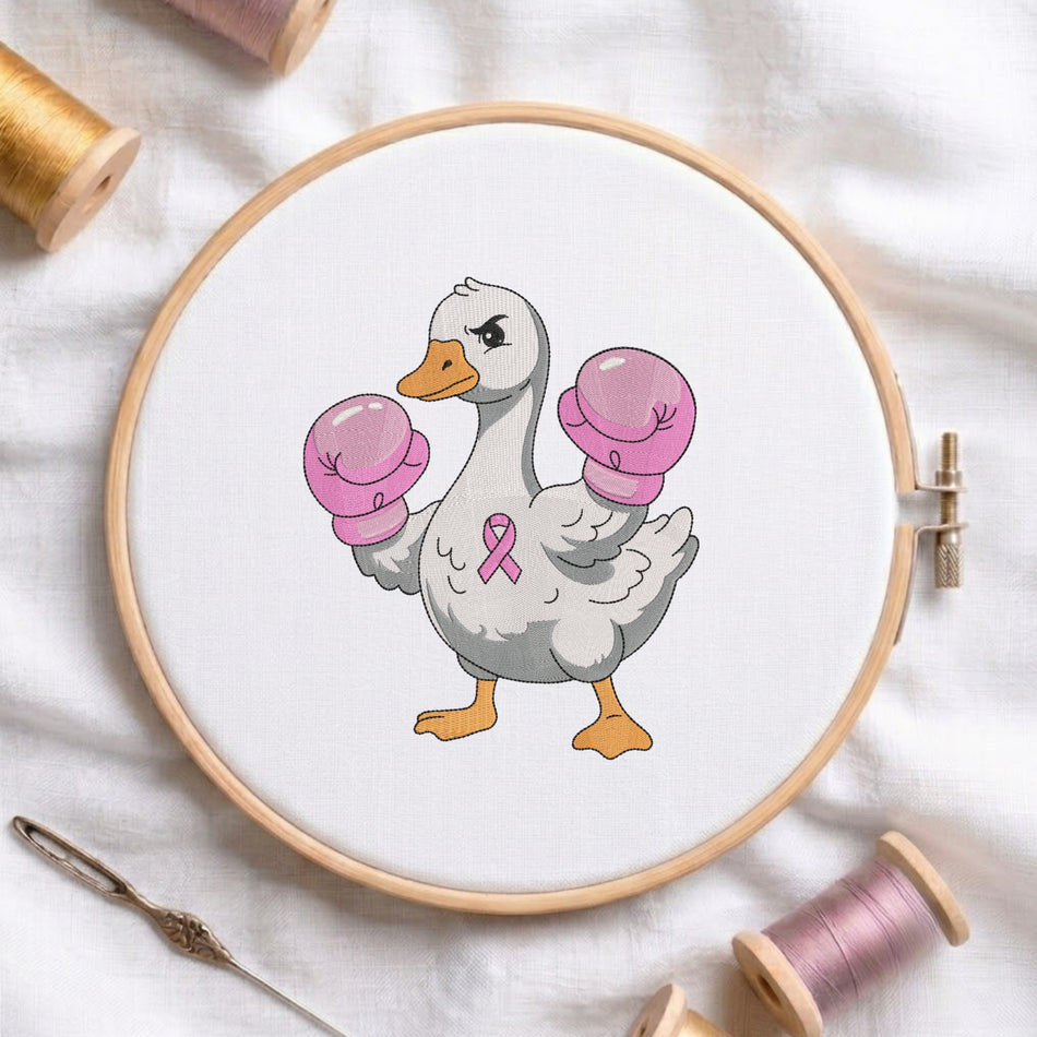 A circular embroidery hoop holding white fabric with a cartoon duck wearing pink boxing gloves and a pink ribbon on its chest. The hoop is styled on a white fabric background with spools of thread and a needle around it.