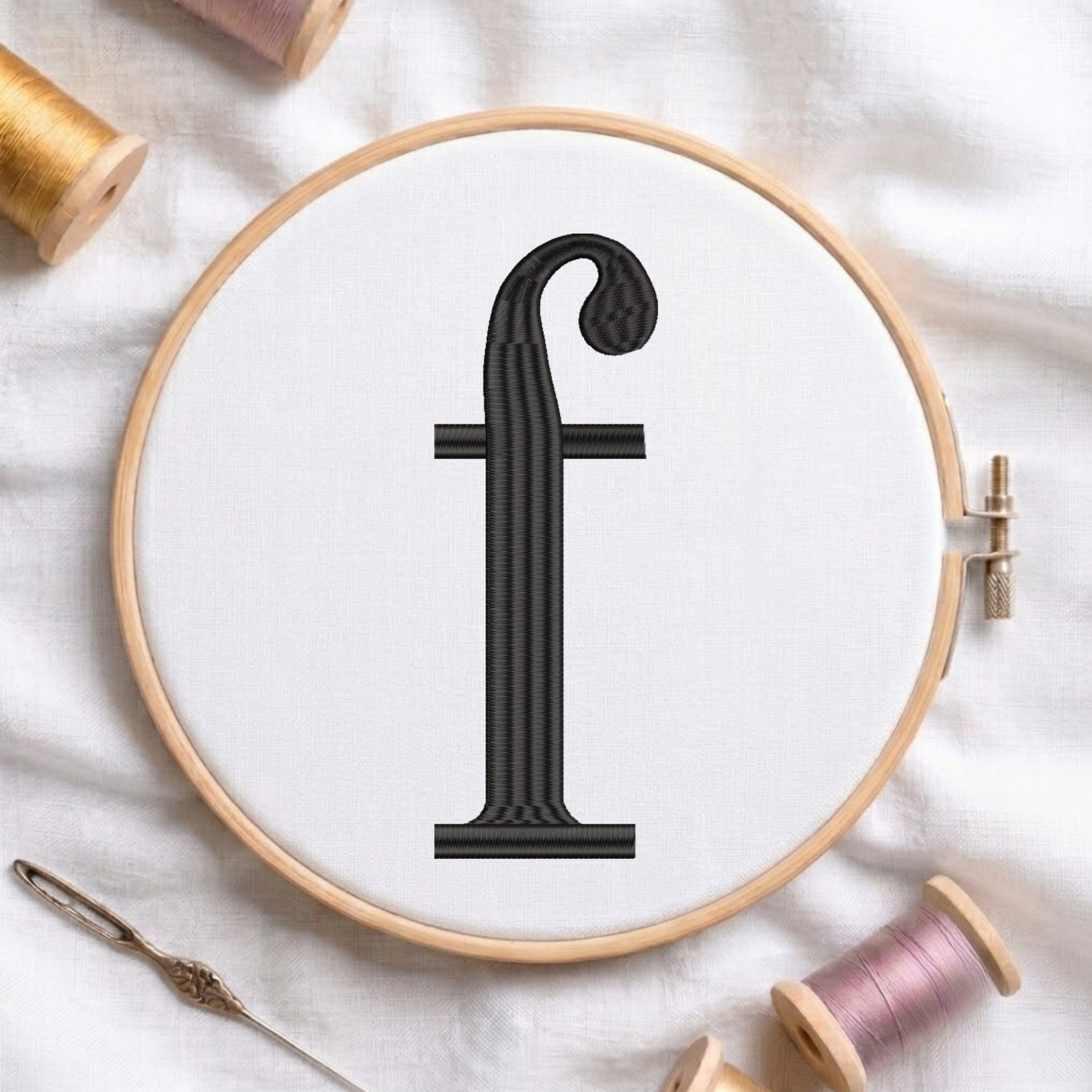 A wooden embroidery hoop holding white fabric with a black embroidered letter 'f' centered. A metal needle lies at the lower left, and spools of thread in gold, pink, and beige are arranged around the hoop on a white fabric background.