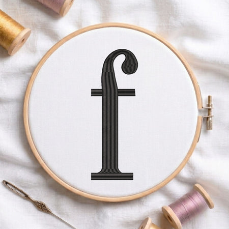 A wooden embroidery hoop holding white fabric with a black embroidered letter 'f' centered. A metal needle lies at the lower left, and spools of thread in gold, pink, and beige are arranged around the hoop on a white fabric background.