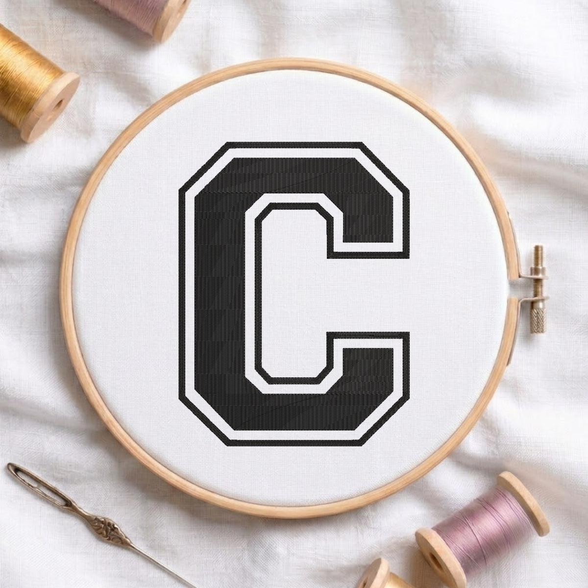 A wooden embroidery hoop holding white fabric with a large black letter C outlined in white, surrounded by spools of thread and a needle on a white fabric background.