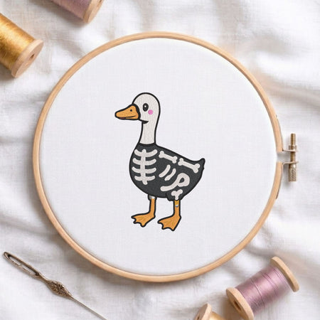 Cute duck dressed in a skeleton costume for Halloween machine embroidery design shown in a wooden embroidery hoop on white fabric with thread spools and a needle   