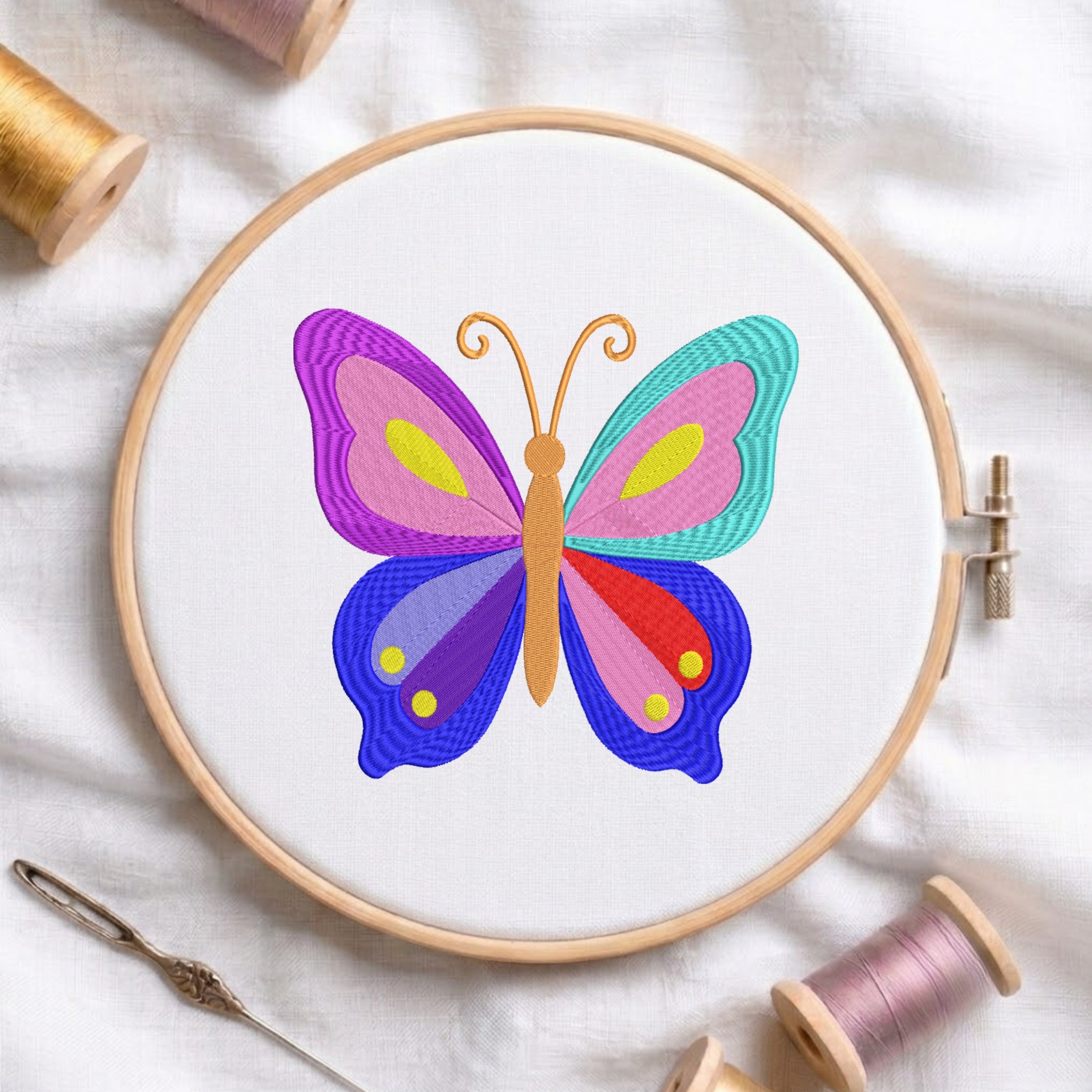 A colorful embroidered butterfly design in a round wooden embroidery hoop, surrounded by spools of thread and a needle on a white fabric background.