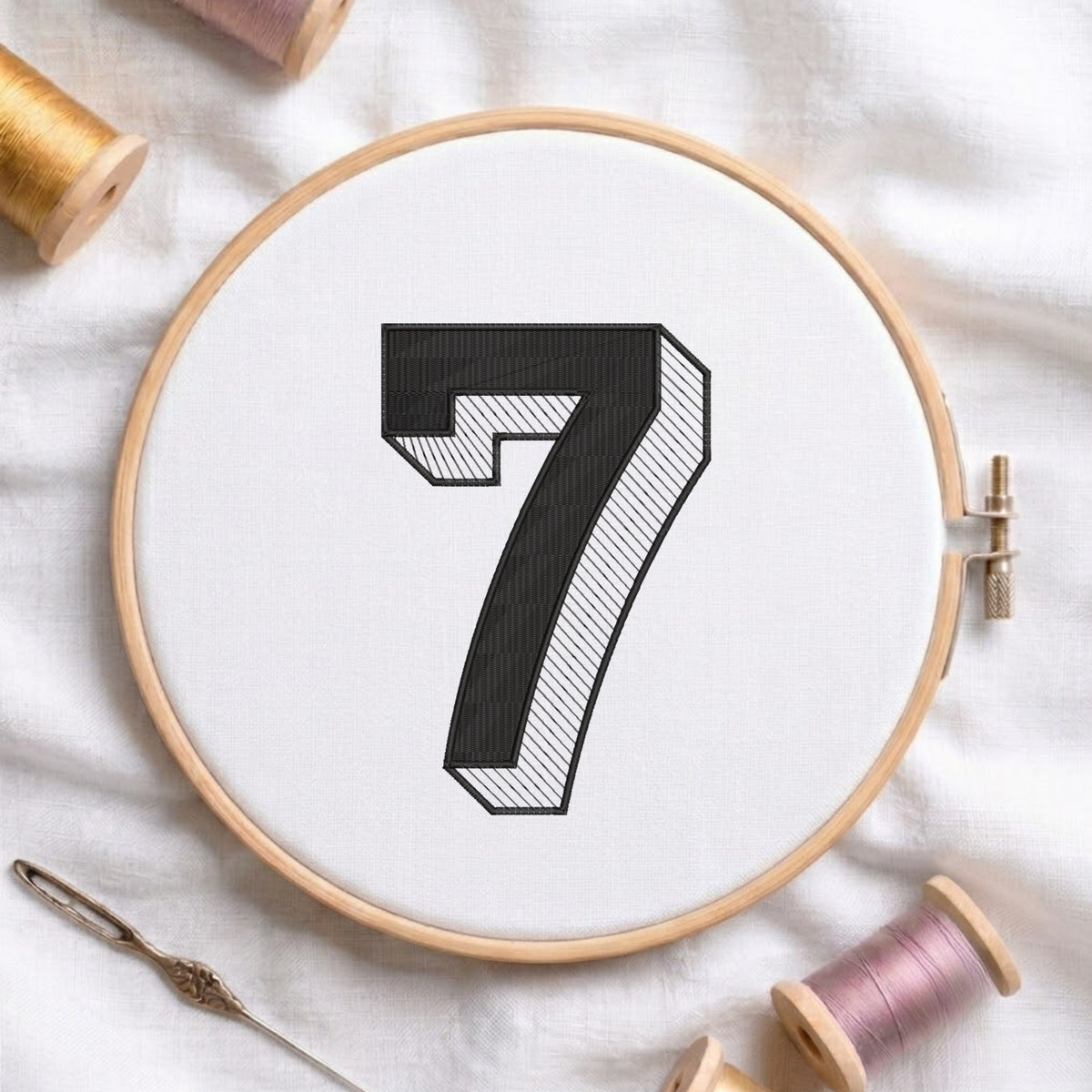 A wooden embroidery hoop holding white fabric with a large black number 7 design, surrounded by spools of thread and a needle on a white fabric background.