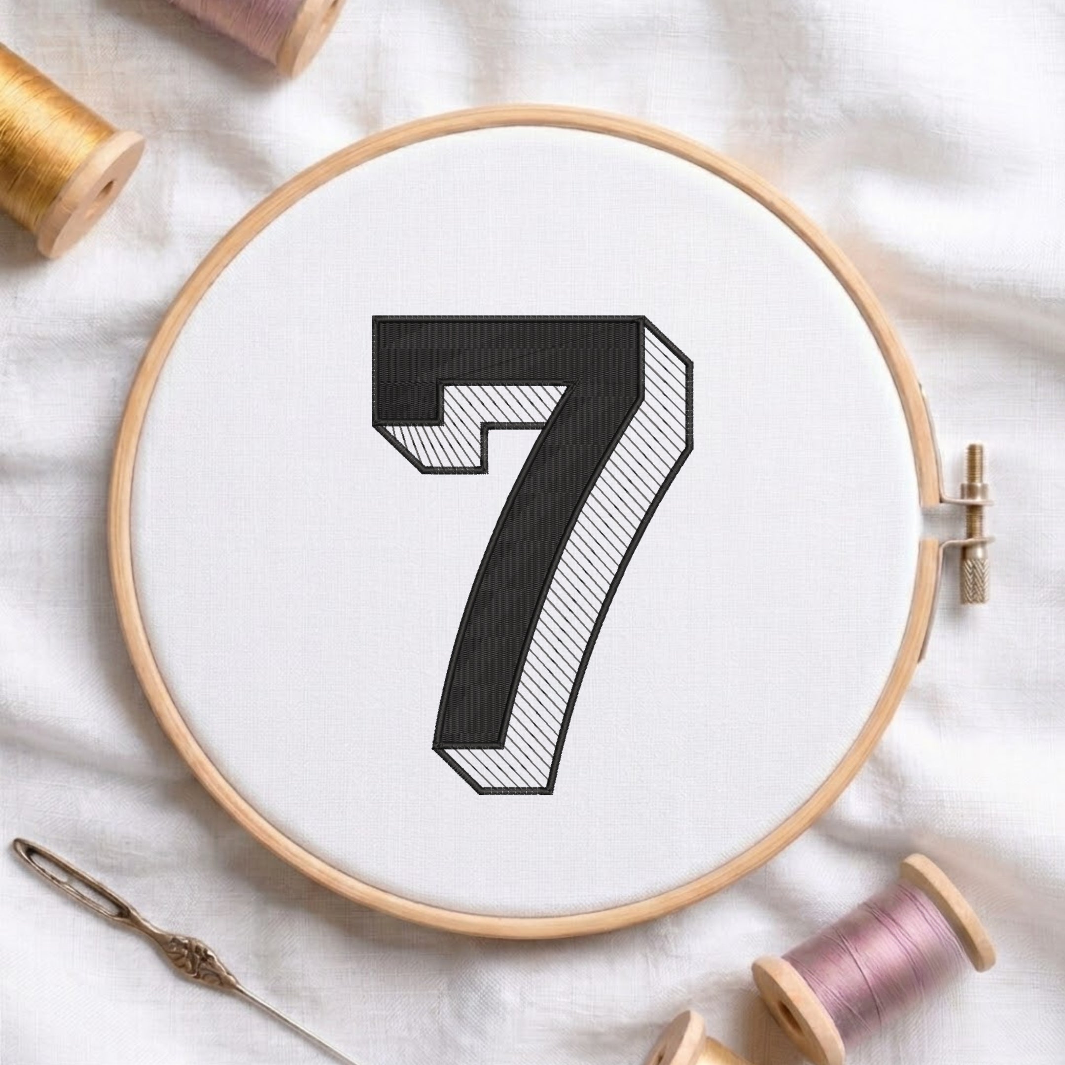 A wooden embroidery hoop holding white fabric with a large black number 7 design, surrounded by spools of thread and a needle on a white fabric background.