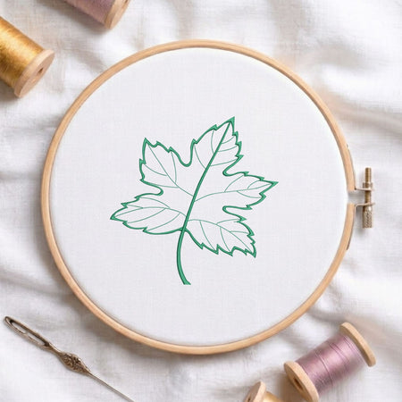 Green maple leaf machine embroidery design displayed in a wooden hoop on white fabric with thread spools and a needle   