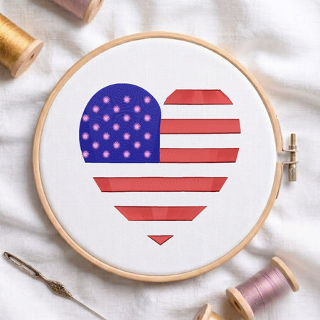 A heart-shaped cross-stitch design featuring the American flag, held in a wooden embroidery hoop on a white fabric background, with spools of thread and a needle around it.
