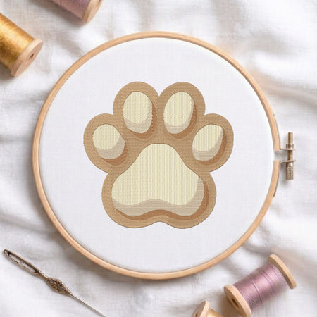 A circular embroidery hoop holding white fabric with a paw print design in layered beige and cream tones. The hoop is styled on a white fabric background with spools of thread and a needle around it.