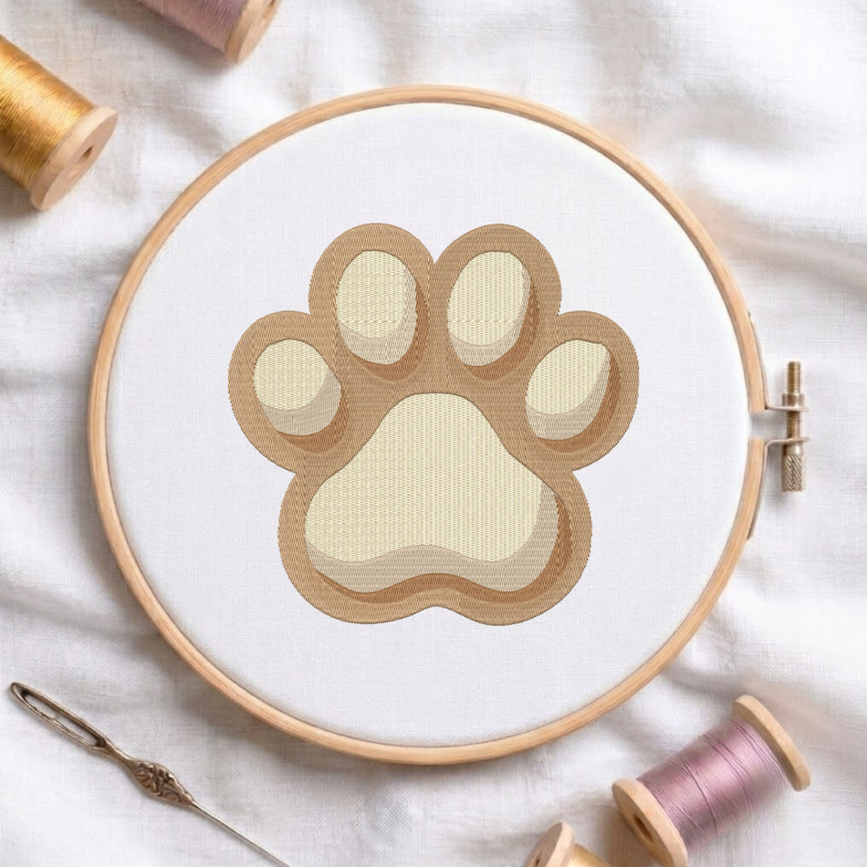 A circular embroidery hoop holding white fabric with a paw print design in layered beige and cream tones. The hoop is styled on a white fabric background with spools of thread and a needle around it.