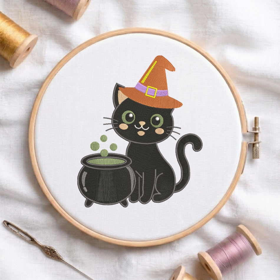 A circular embroidery hoop holding white fabric with a cartoon black cat wearing an orange witch hat and sitting beside a black cauldron with green liquid. The hoop is styled on a white fabric background with spools of thread and a needle around it.