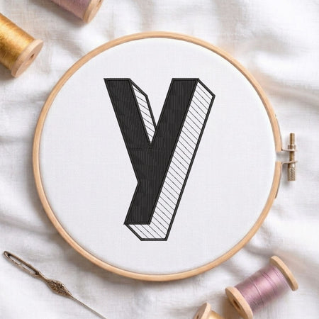 A wooden embroidery hoop holding white fabric with a large black letter 'Y' design, surrounded by embroidery thread spools and a needle on a white fabric background.
