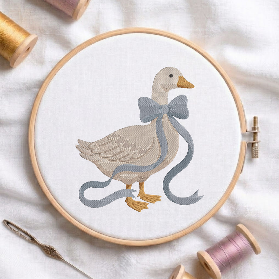 Cute goose wearing a blue ribbon bow machine embroidery design displayed inside a round wooden hoop on white linen fabric, accompanied by wooden thread spools and a needle.