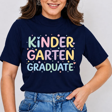 A woman wearing a navy blue t-shirt with a colorful graphic that reads 'KINDER GARTEN GRADUATE' in pastel letters, paired with blue jeans against a light gray background.