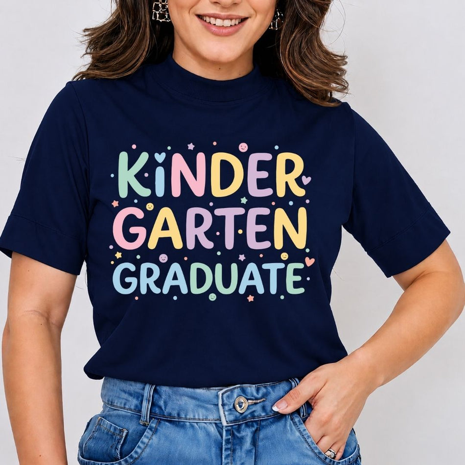 A woman wearing a navy blue t-shirt with a colorful graphic that reads 'KINDER GARTEN GRADUATE' in pastel letters, paired with blue jeans against a light gray background.