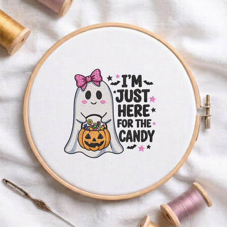  "I'm Just Here for the Candy" cute ghost holding a pumpkin bucket with bats, stars, and a pink polka dot bow machine embroidery design displayed in a wooden embroidery hoop on white fabric with thread spools and a needle   