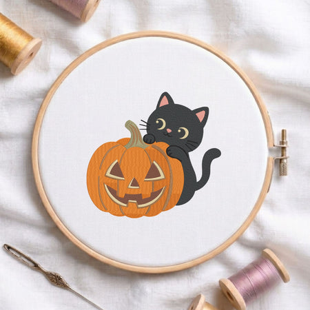 Cute black cat peeking behind a carved jack-o-lantern pumpkin Halloween machine embroidery design shown in a wooden embroidery hoop on white fabric with thread spools and a needle