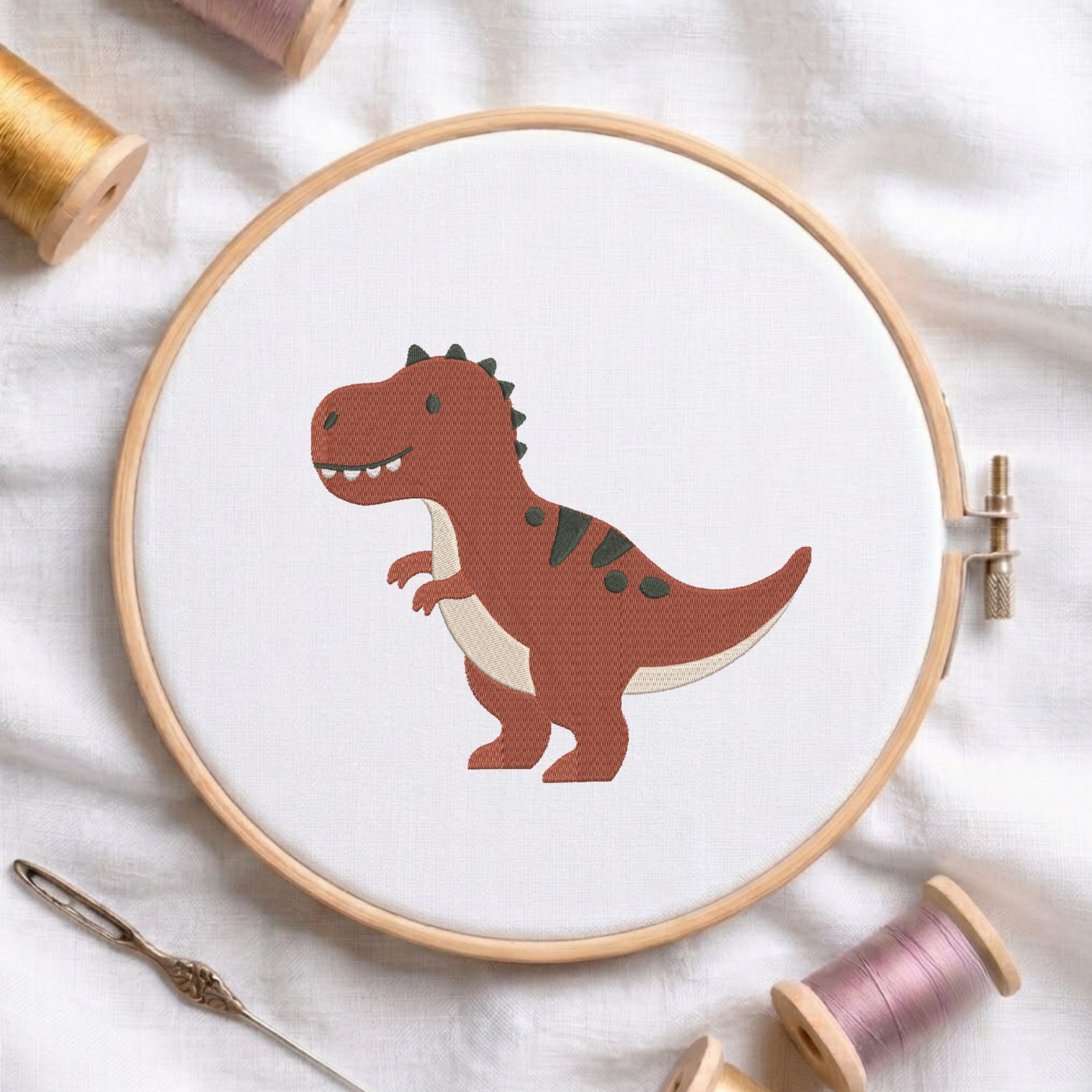 A wooden embroidery hoop on white fabric, featuring a cartoon-style brown dinosaur with green spots and a cream belly. Spools of thread, a needle, and a pin are arranged around the hoop.