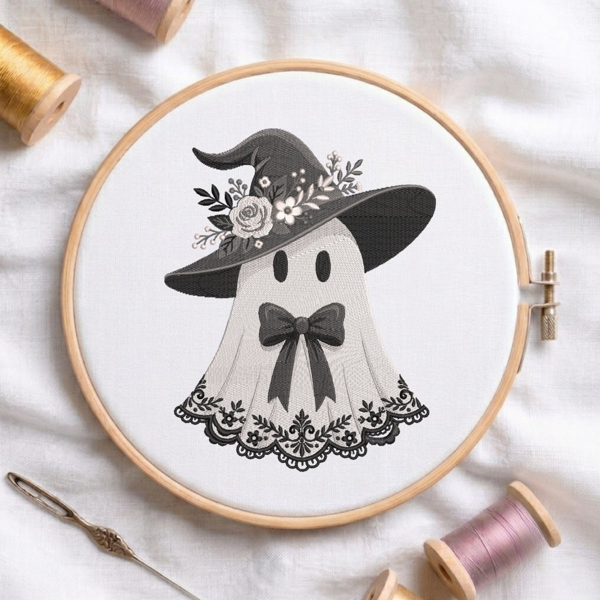 A circular embroidery hoop holding white fabric with a black-and-white stitched design of a ghost wearing a witch hat with flowers, surrounded by spools of thread and a needle on a white fabric background.