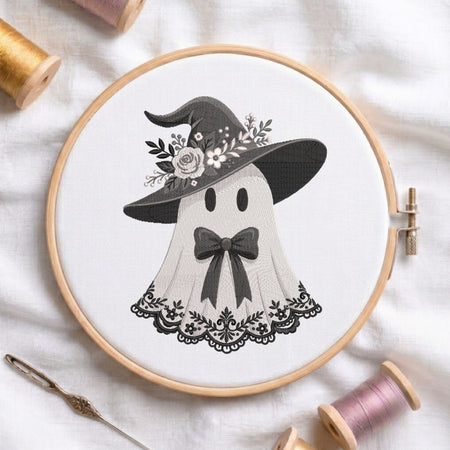 A circular embroidery hoop holding white fabric with a black-and-white stitched design of a ghost wearing a witch hat with flowers, surrounded by spools of thread and a needle on a white fabric background.