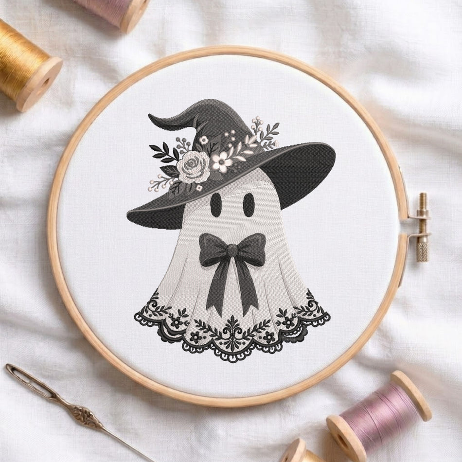 A circular embroidery hoop holding white fabric with a black-and-white stitched design of a ghost wearing a witch hat with flowers, surrounded by spools of thread and a needle on a white fabric background.