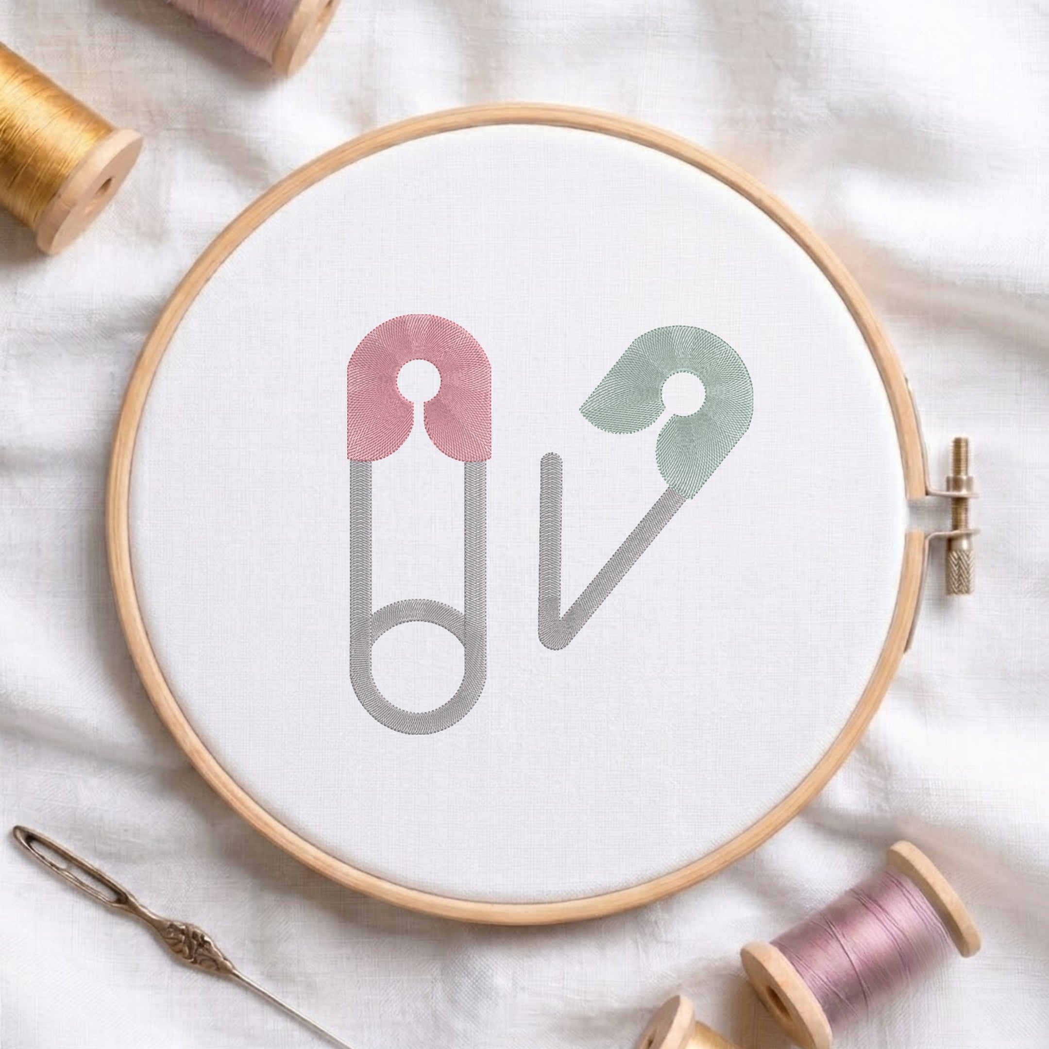 A circular embroidery hoop holding white fabric with a minimalist design of two safety pins, one in dusty pink and one in sage green, both outlined in gray. The hoop is styled on a white fabric background with spools of thread and a needle.