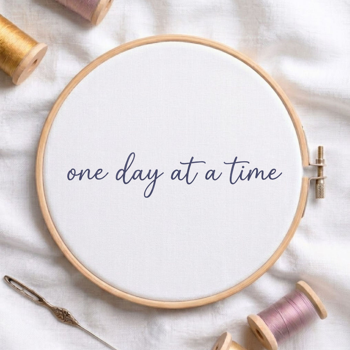 A round embroidery hoop holding white fabric with the phrase “one day at a time” stitched in navy script. The hoop is styled on a white fabric background with spools of thread and a needle around it.