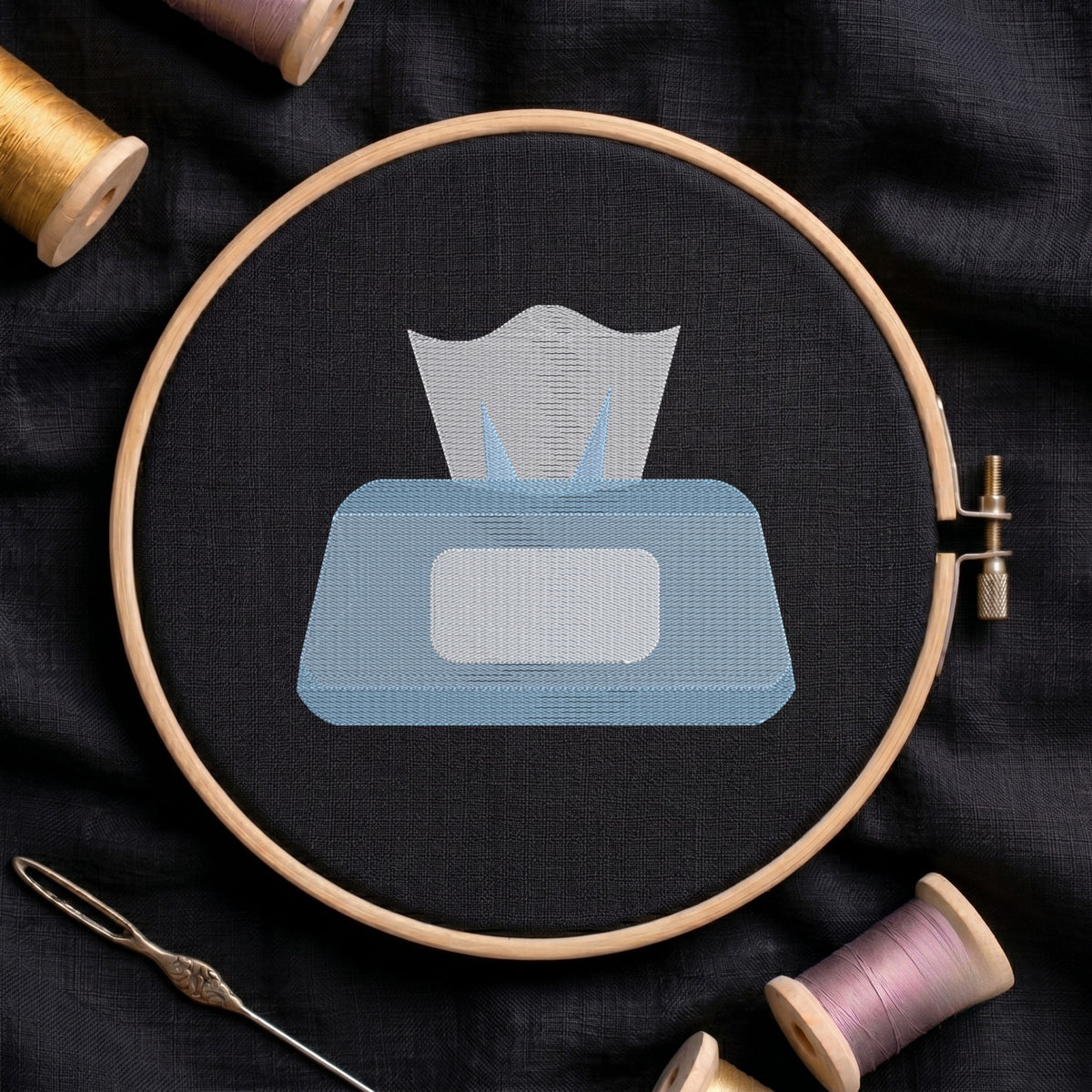 A circular embroidery hoop holding black fabric with a stitched tissue box design in light blue and white, surrounded by spools of thread and a needle on a dark fabric background.
