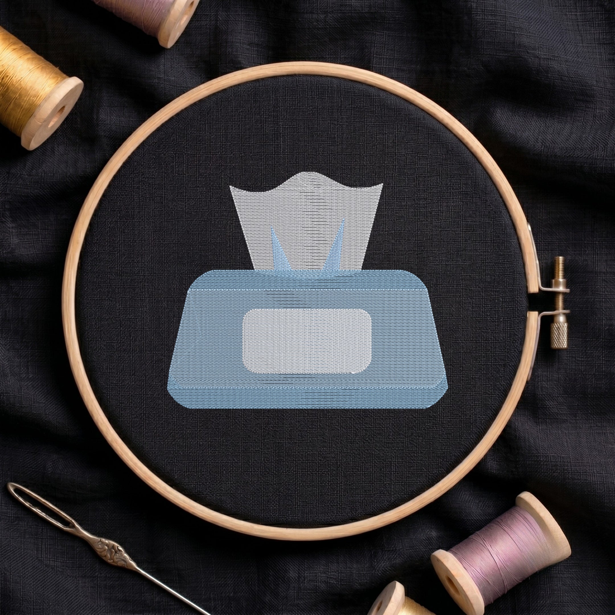 A circular embroidery hoop holding black fabric with a stitched tissue box design in light blue and white, surrounded by spools of thread and a needle on a dark fabric background.