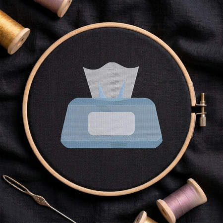 A circular embroidery hoop holding black fabric with a stitched tissue box design in light blue and white, surrounded by spools of thread and a needle on a dark fabric background.