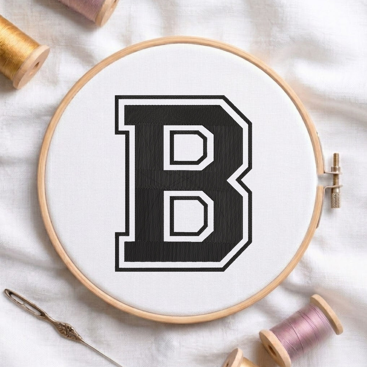 A wooden embroidery hoop holding white fabric with a large black letter B outlined in white, surrounded by spools of thread and a needle on a white fabric background.