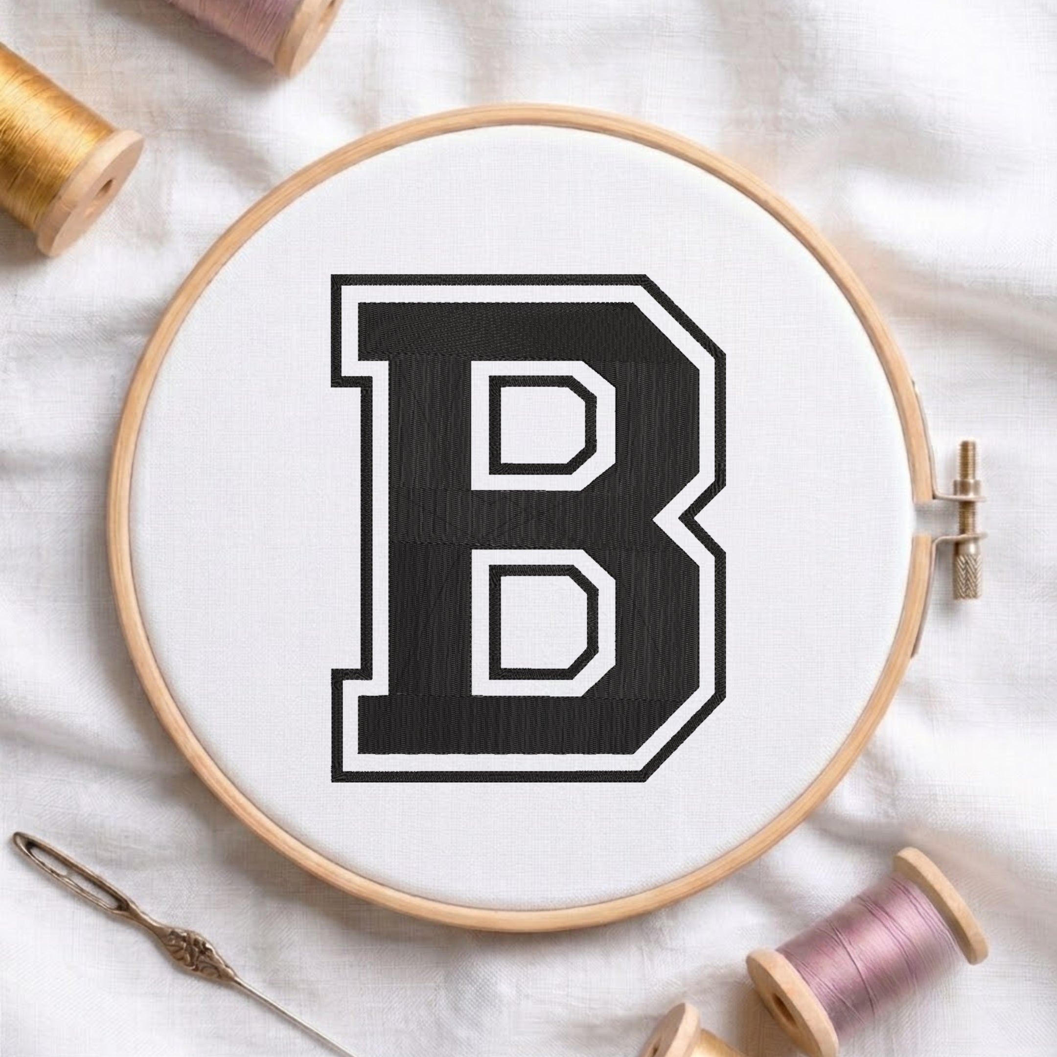 A wooden embroidery hoop holding white fabric with a large black letter B outlined in white, surrounded by spools of thread and a needle on a white fabric background.