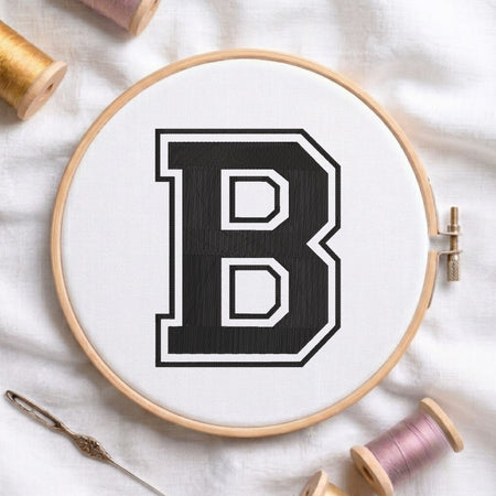 A wooden embroidery hoop holding white fabric with a large black letter B outlined in white, surrounded by spools of thread and a needle on a white fabric background.