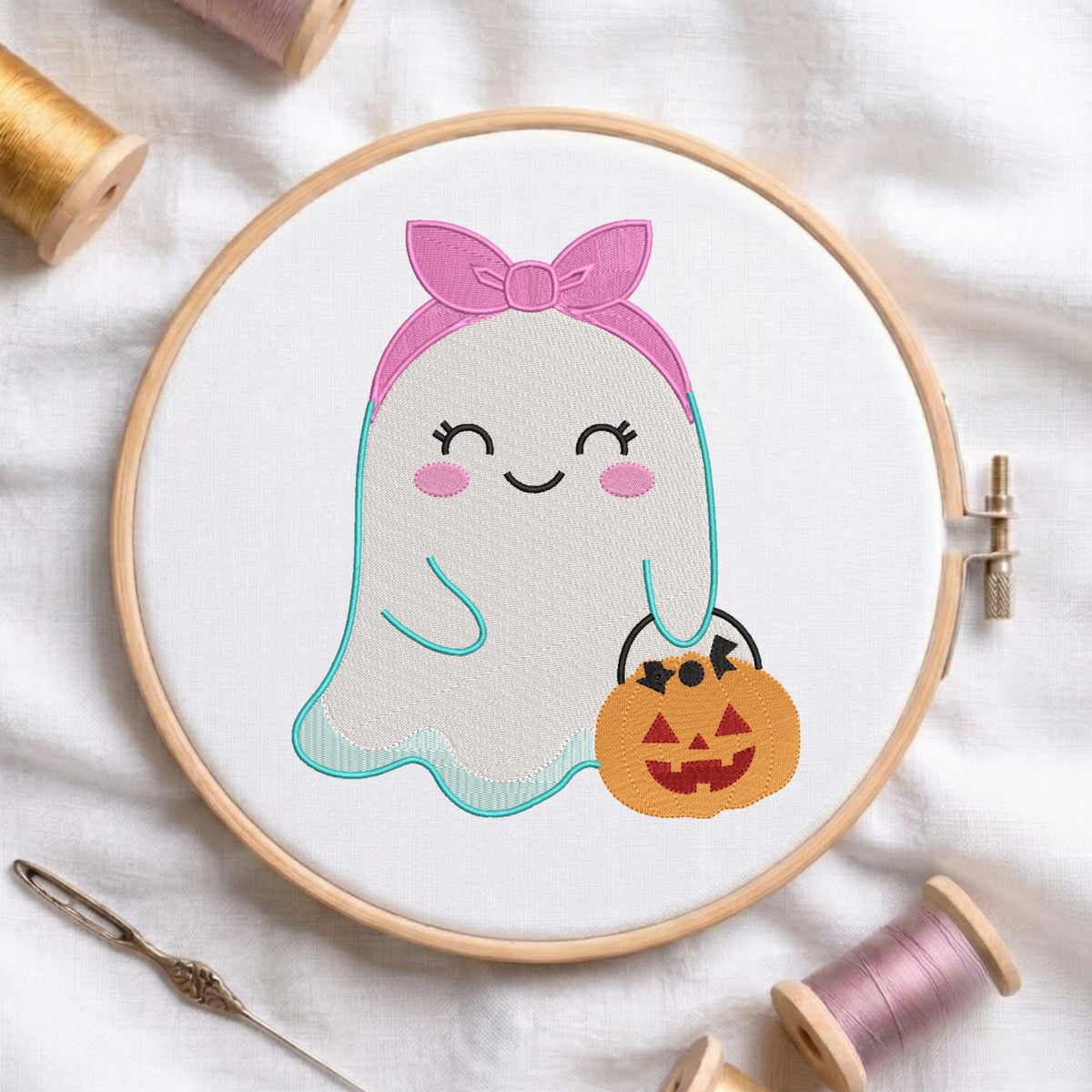 A circular embroidery hoop holding white fabric with a cute embroidered ghost character wearing a pink bow and holding a jack-o'-lantern bucket, surrounded by spools of thread and a needle on a white fabric background.