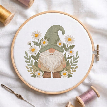 A circular embroidery hoop holding white fabric with a stitched gnomes design surrounded by daisies and green leaves. The hoop is styled on a white fabric background with spools of thread, a needle, and wooden pegs.