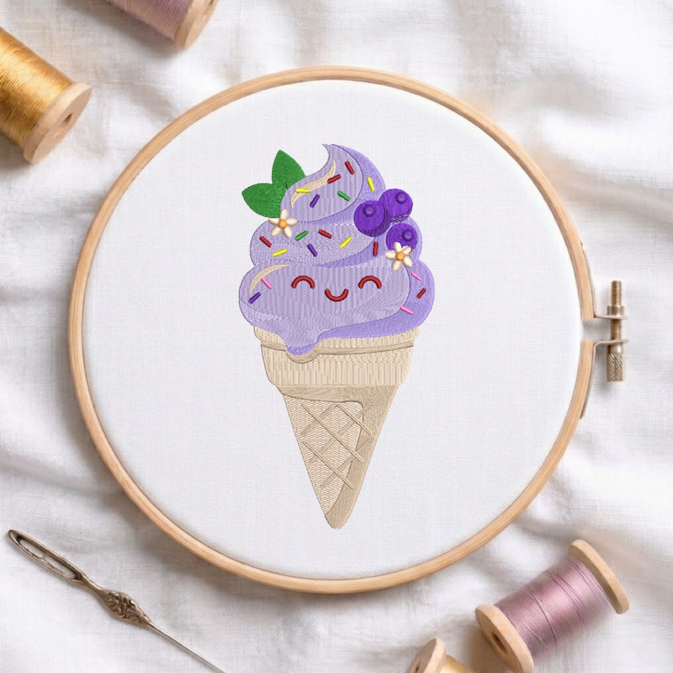 A circular embroidery hoop holds a white fabric with an embroidered ice cream cone design. The swirl is lavender purple with small multicolored sprinkles, two purple berries, and a green leaf, set in a beige waffle cone.