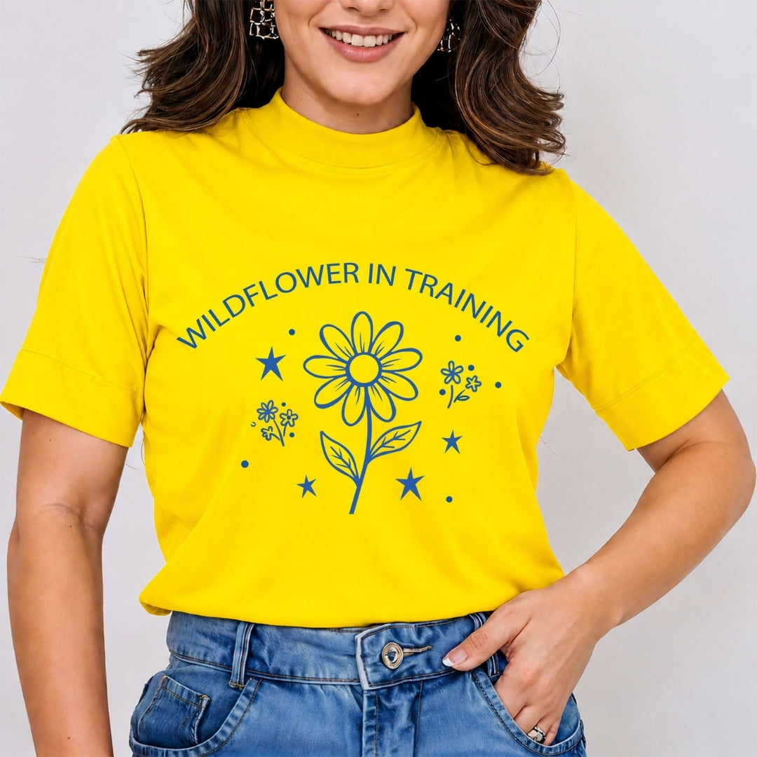 Woman wearing a bright yellow mock neck t-shirt featuring a blue retro line art "Wildflower in Training" graphic print with a daisy flower and stars, tucked into blue jeans.