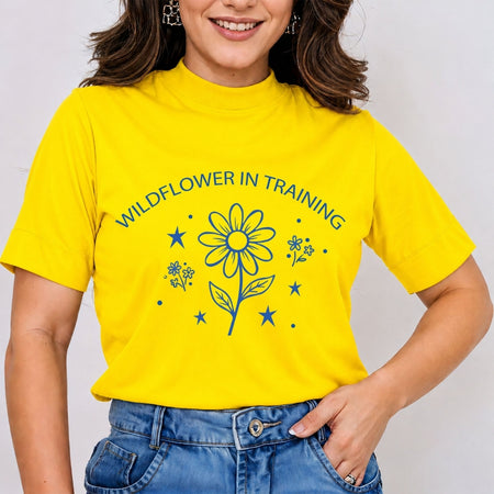 Woman wearing a bright yellow mock neck t-shirt featuring a blue retro line art "Wildflower in Training" graphic print with a daisy flower and stars, tucked into blue jeans.