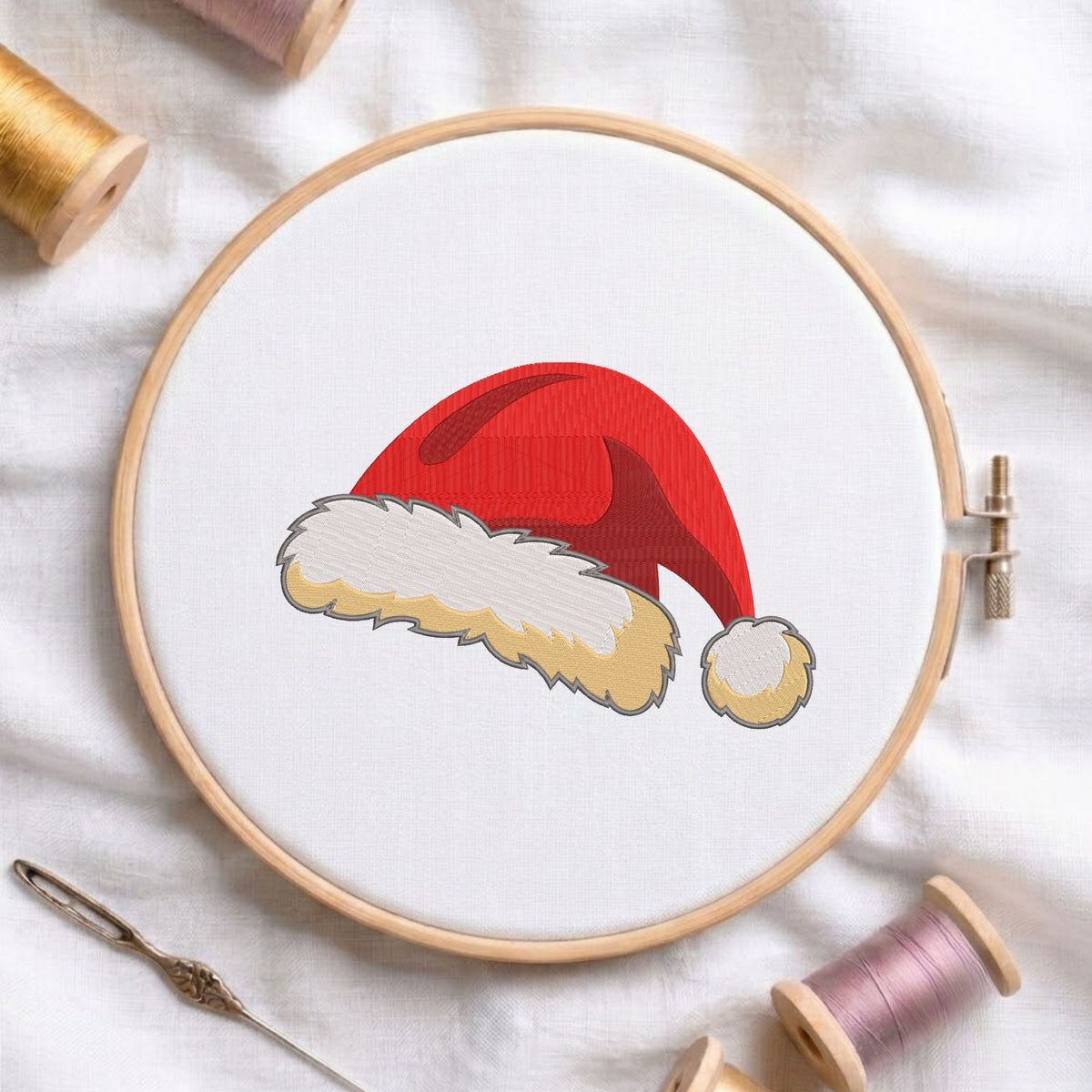 Santa Claus hat machine embroidery design with bright red fabric and fluffy white trim displayed inside a round wooden hoop on white linen fabric, accompanied by wooden thread spools and a needle.