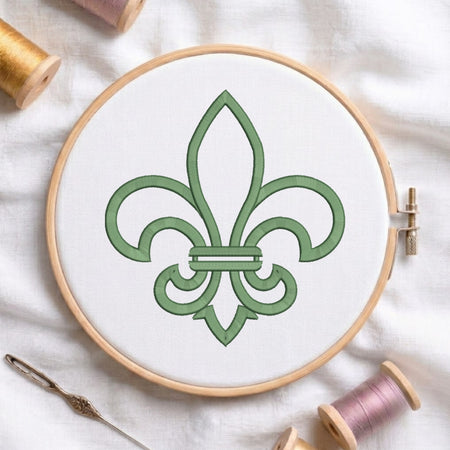 A circular embroidery hoop holding white fabric with a green fleur-de-lis design, surrounded by spools of thread and a needle on a white fabric background.