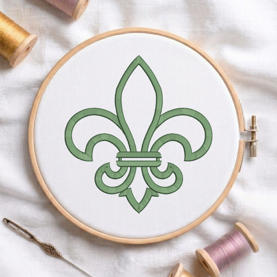 A circular embroidery hoop holding white fabric with a green fleur-de-lis design, surrounded by spools of thread and a needle on a white fabric background.