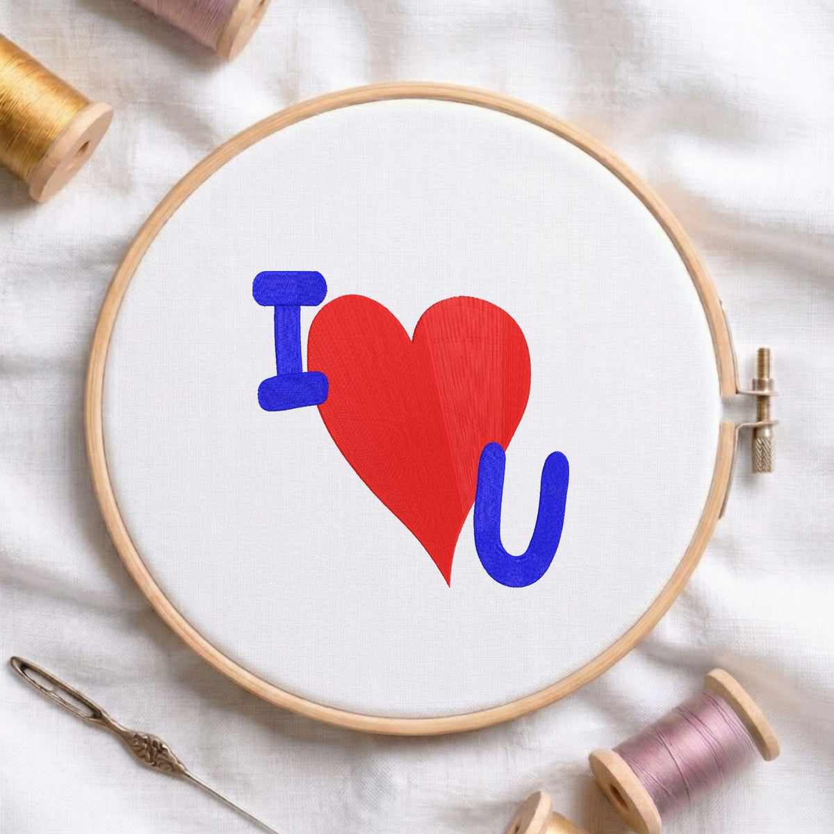 I Love You" text and heart machine embroidery design displayed in a wooden hoop on white fabric with thread spools and a needle