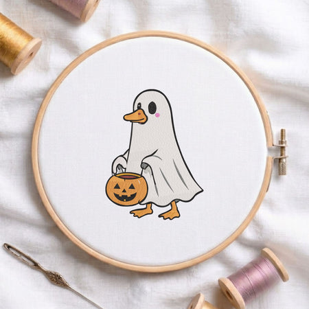 Cute duck dressed in a ghost costume holding a jack-o-lantern trick-or-treat bucket Halloween machine embroidery design shown in a wooden embroidery hoop on white fabric with thread spools and a needle