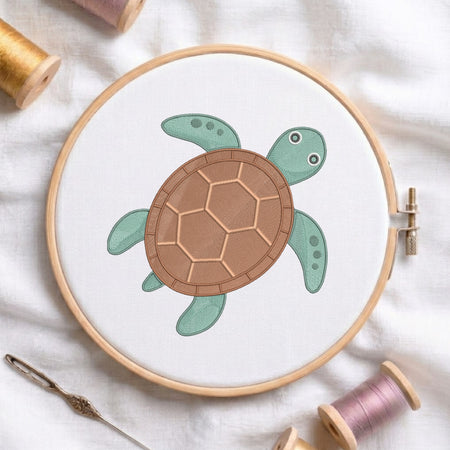 A wooden embroidery hoop holds white fabric with a stylized sea turtle design in mint green and brown. The hoop is photographed on a white fabric surface with spools of thread and a needle around it.