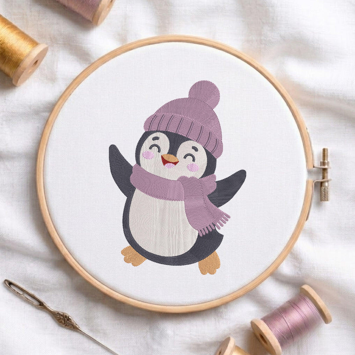 A circular embroidery hoop holds white fabric with a stitched cartoon penguin wearing a lavender knit hat and matching scarf. The hoop is styled on a white fabric background with spools of thread and a needle around it.