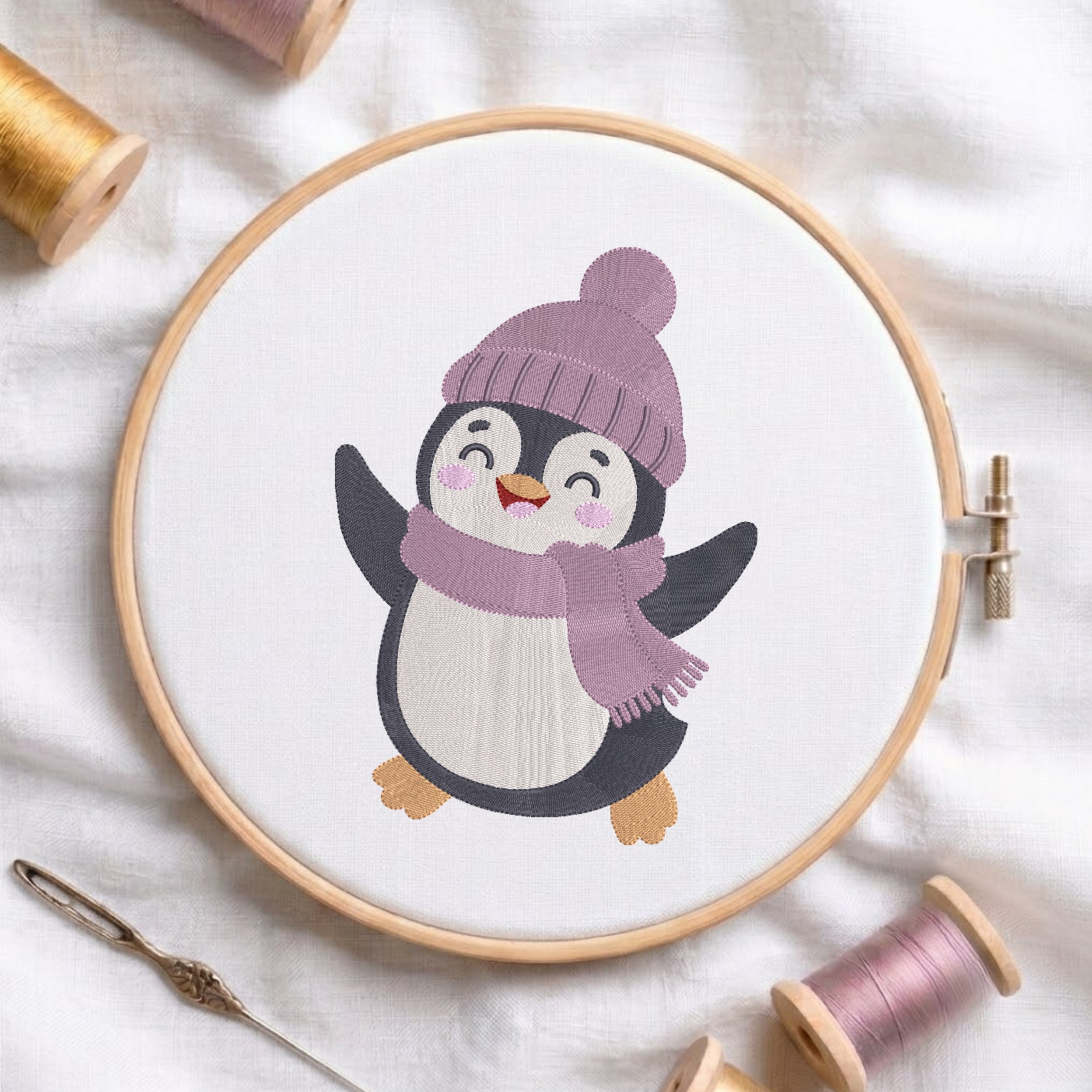 A circular embroidery hoop holds white fabric with a stitched cartoon penguin wearing a lavender knit hat and matching scarf. The hoop is styled on a white fabric background with spools of thread and a needle around it.