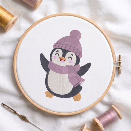 A circular embroidery hoop holds white fabric with a stitched cartoon penguin wearing a lavender knit hat and matching scarf. The hoop is styled on a white fabric background with spools of thread and a needle around it.